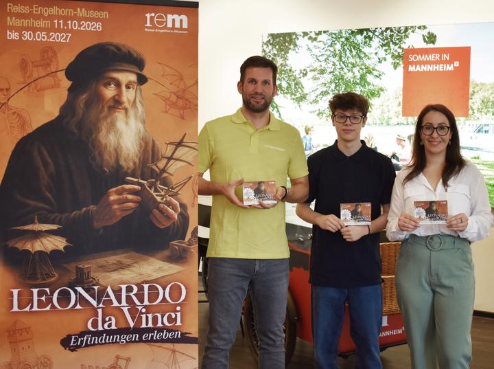 Links ist der Aufsteller zu einer Ausstellung mit der Darstellung von Leonardo da Vinci zu sehen. Rechts daneben stehen zwei Männer und eine Frau, die kleine Faltkarten mit Ausflugstipps vor sich halten.