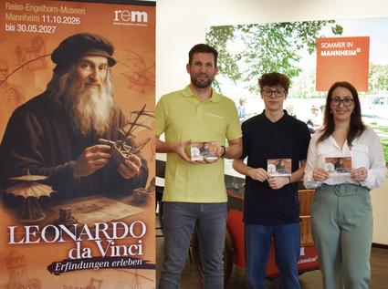 Präsentation der neuen Faltkarte in der Tourist Information Mannheim Links ist der Aufsteller zu einer Ausstellung mit der Darstellung von Leonardo da Vinci zu sehen. Rechts daneben stehen zwei Männer und eine Frau, die kleine Faltkarten mit Ausflugstipps vor sich halten.