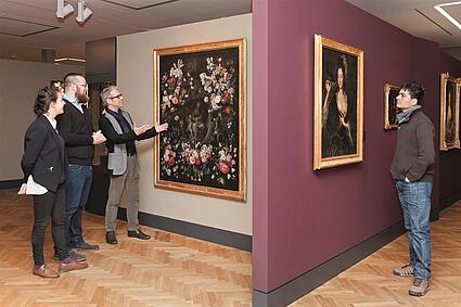 Vier Personen betrachten in einer Ausstellung Gemälde. Auf dem einen Gemälde sind zahlreiche Blüten und auf dem anderen eine Frau zu sehen. 