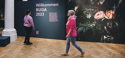 Zwei Besucherinnen vor Wand mit Blumentapete und Schriftzug "Willkommen BUGA 2023"