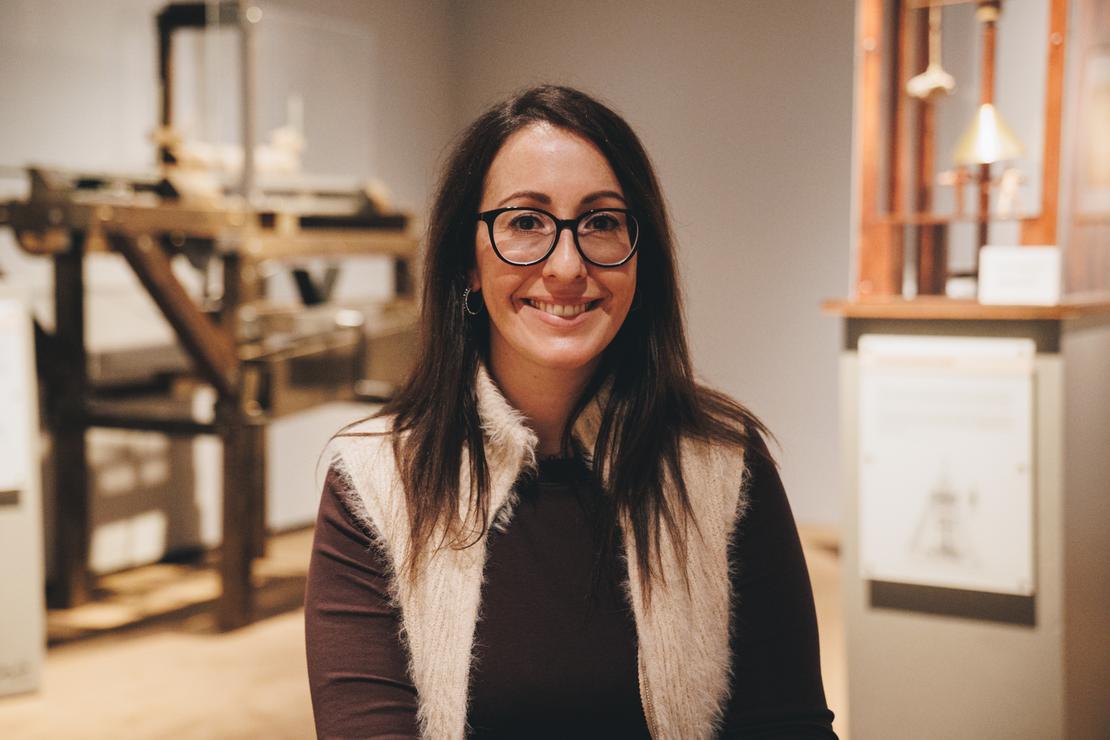 Kuratorin Giulia Worf Kuratorin Giulia Worf sitzt in einer Ausstellungsfläche. Sie hat lange dunkle Haare und trägt eine Brille, einen dunkelbraunen Pulli und eine heller Weste. Im Hintergrund sind Mitmach-Stationen zu erkennen.