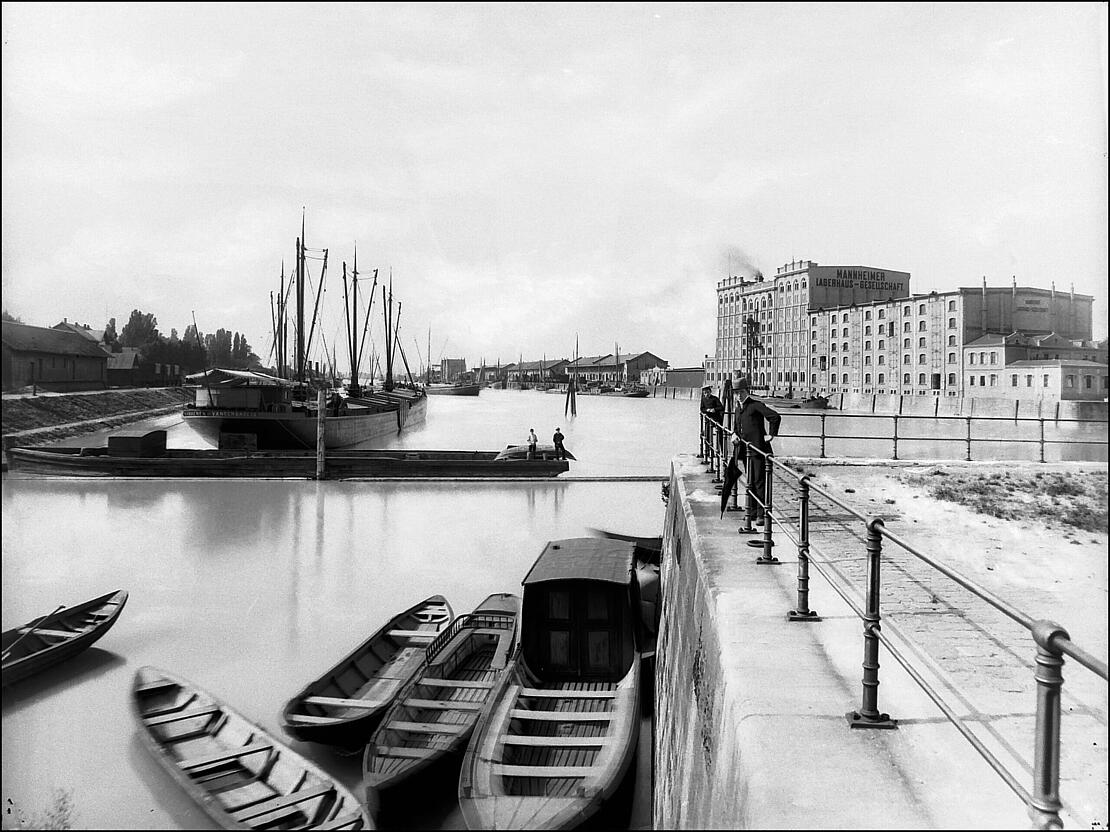 Historische Fotografie mit Blick in Hafenbecken, vorne liegen drei Holzboote, rechts an der Kaimauer lehnt ein Mann mit Hut und zugeklapptem Regenschirm