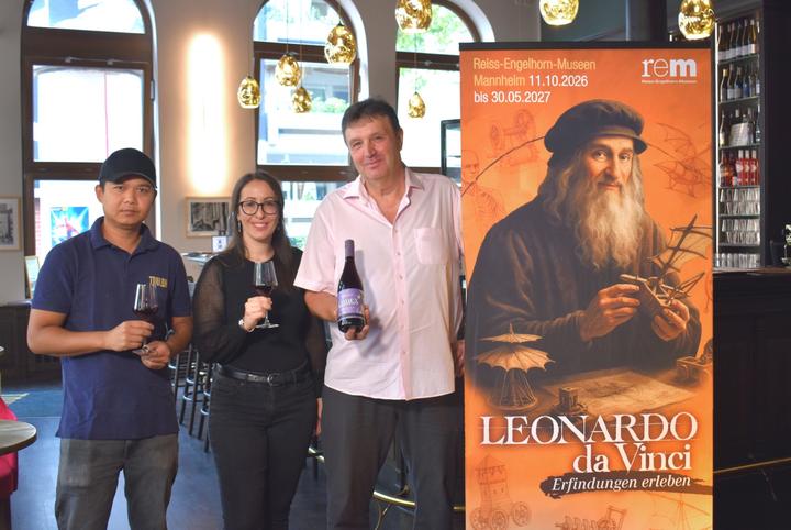Wein zur Leonardo-Ausstellung Drei Personen mit weingläsern in der Hand stehen in einem Lokal, rechts von ihnen stecht ein Aufsteller mit dem Plakat zur Ausstellung "Leonardo da Vinci - Erfindungen erleben"