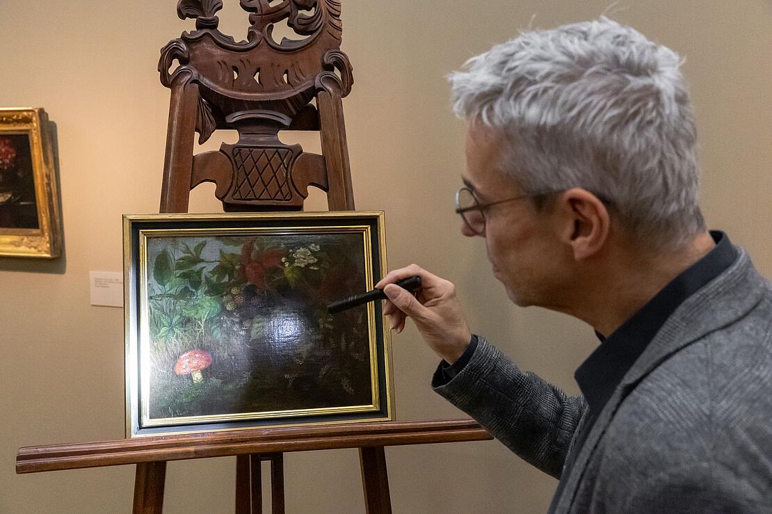 Ein Ölbild mit Waldstillleben steht auf einer Staffelei, Mann leuchtet Detail an