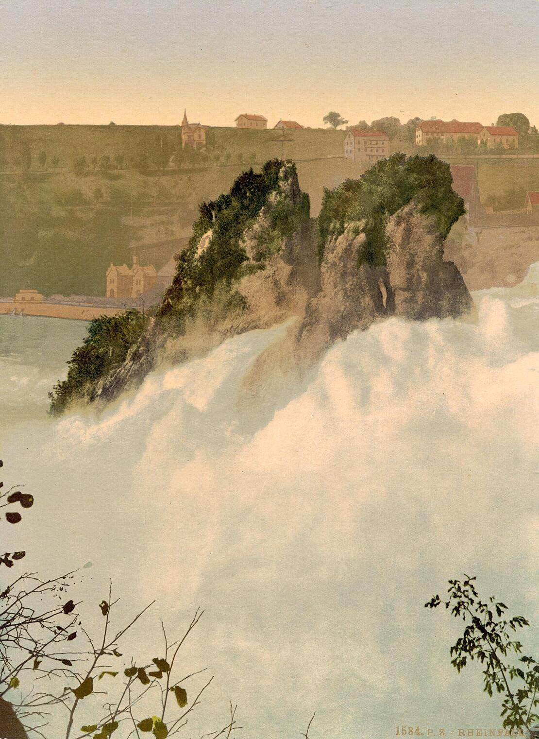 Historische Farbfotografie zeigt den Rheinfall bei Schaffhausen: Zwei Berggipfel schauen aus gischspritzendem Wasser hervor, im Hintergrund ist das Flussufer zu sehen