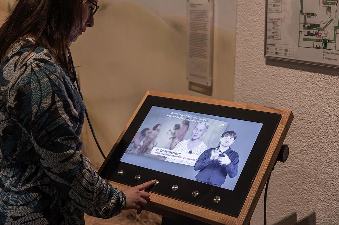Frau drückt einen Knopf an einem Monitor, auf dem ein video mit Gebärdendolmetscher zu sehen ist.