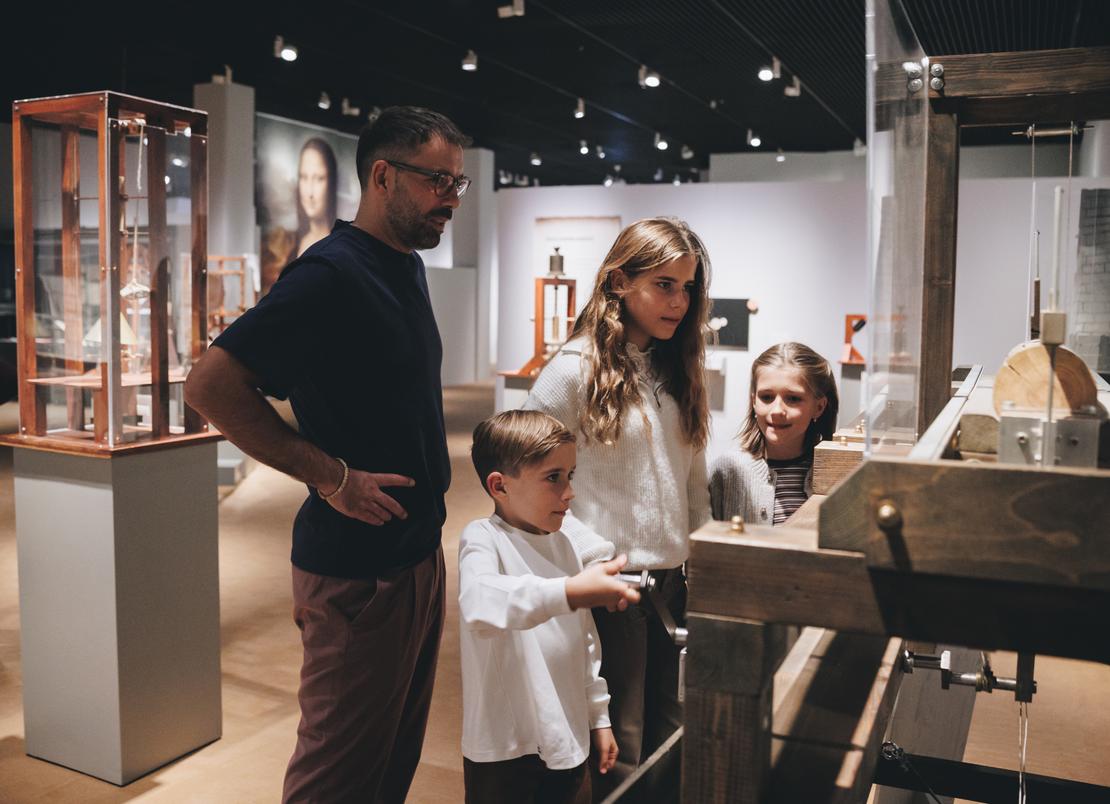Mitmach-Stationen sorgen in der Ausstellung für Bewegung Ein Mann und drei Kinder testen in einem Ausstellungsraum eine Mitmach-Station, mit der man Holzbretter schneiden kann.