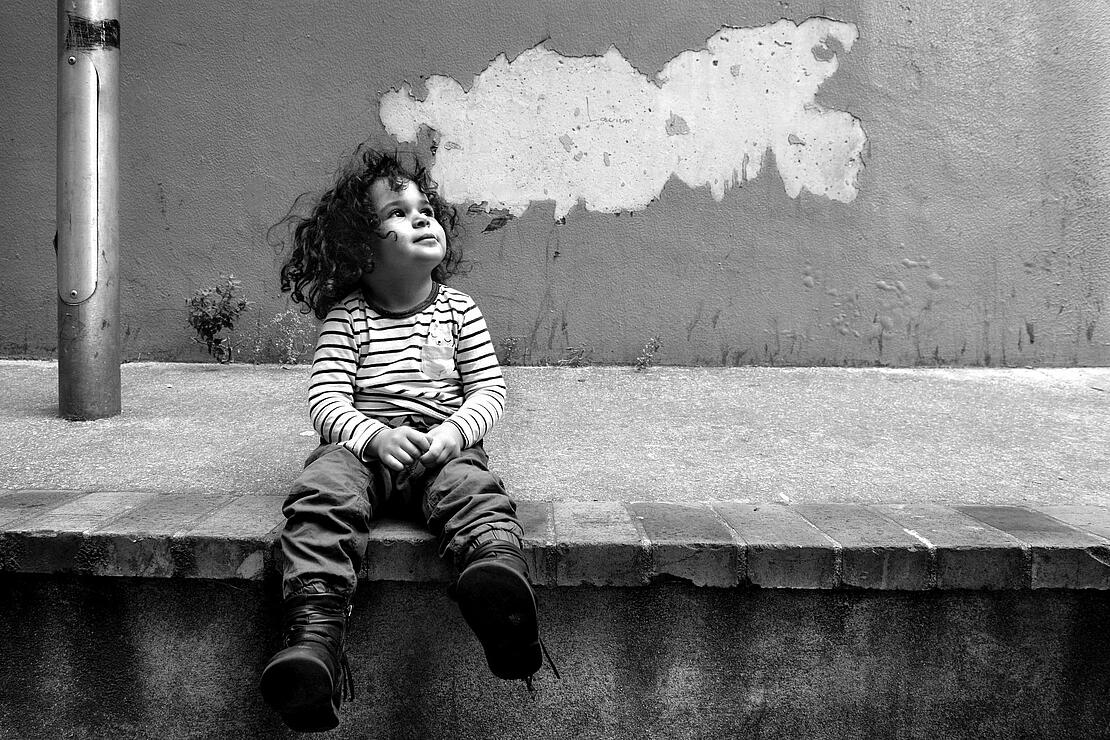 Schwarz-weiß-Bild zeigt ein kleines Mädchen mit dunklen Locken und gestreiftem Shirt, das auf einer Straßenerhebung sitzt und nach oben schaut, hinter ihr eine Wand, an der der Putz abgeplatzt ist