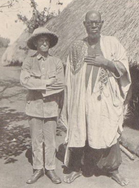 Historische Fotografie zeigt links weißen Mann mit Tropenhelm und rechts daneben größeren farbigen Mann, dahinter strohgedeckte Hütten