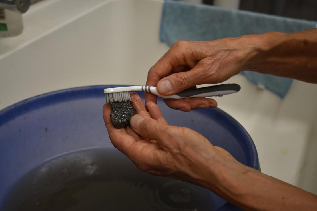 Reinigung der Funde Detailaufnahme auf zwei Hände, die mit einer Zahnbürste einen kleinen schwarzen Gegenstand reinigen, darunter befindet sich eine blaue Wanne mit Wasser.