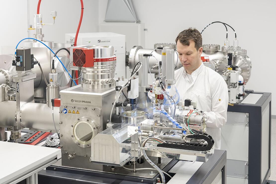 Mann im Laborkittel steht hinter einem großen kompliziert aussehenden Apparat mit vielen Teilen und Schläuchen