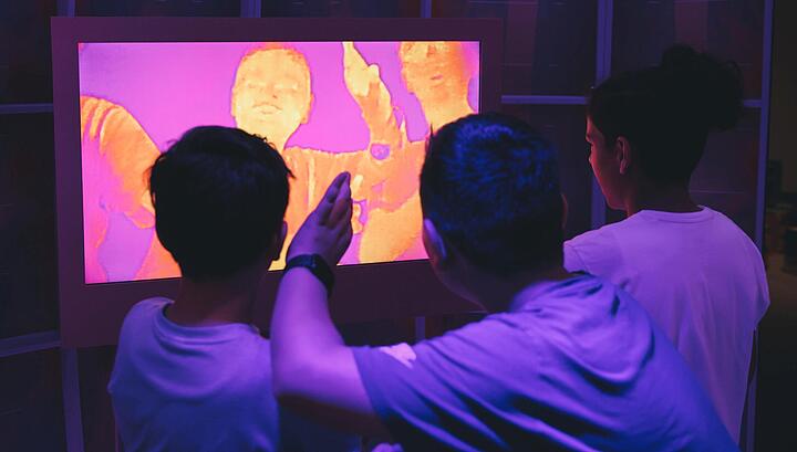 Drei jungen stehen vor einem Monitor, dort sind sie durch eine Wärmebildkamera in Gelbtönen zu sehen