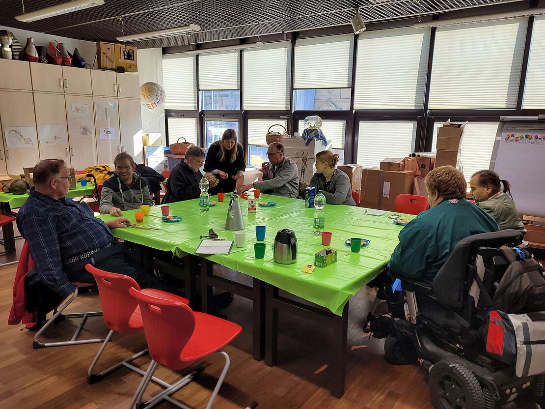 Mehrere Personen mit und ohne Rollstuhl sitzen um einen Tisch mit grüner Tischdecke