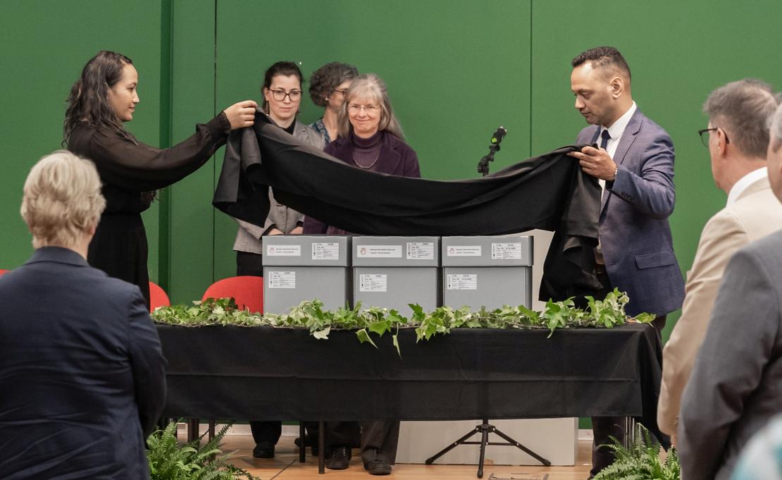 Übergabezeremonie der toi moko Auf einem Tisch befinden sich drei graue Kartons. Eine Frau und ein Mann decken die Kartons mit einem schwarzen Tuch ab. Hinter und vor dem Tisch sind weitere Personen zu sehen.