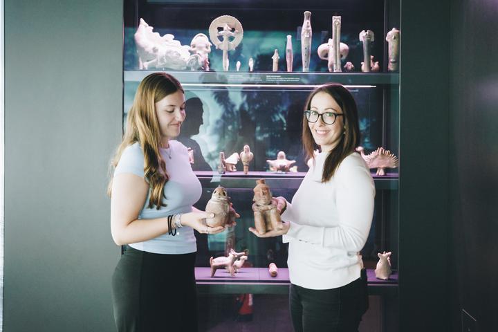 Blick in die Ausstellung „MusikWelten“ Zwei Frauen stehen in einem abgedunkelten Ausstellungsraum vor einer Vitrine mit Tonobjekten. Sie halten jeweils ein Objekt in den Händen.