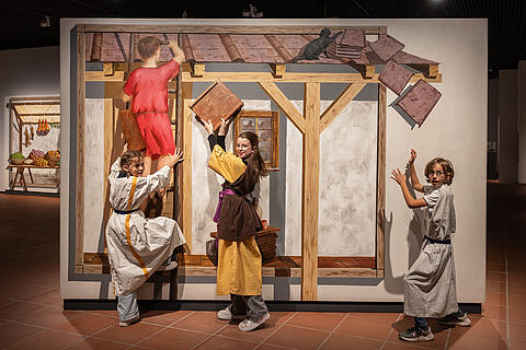 Kinder mit Tuniken als Römer verkleidet setzen sich vor einer Kulisse in Szene, die eine römische Baustelle zeigt