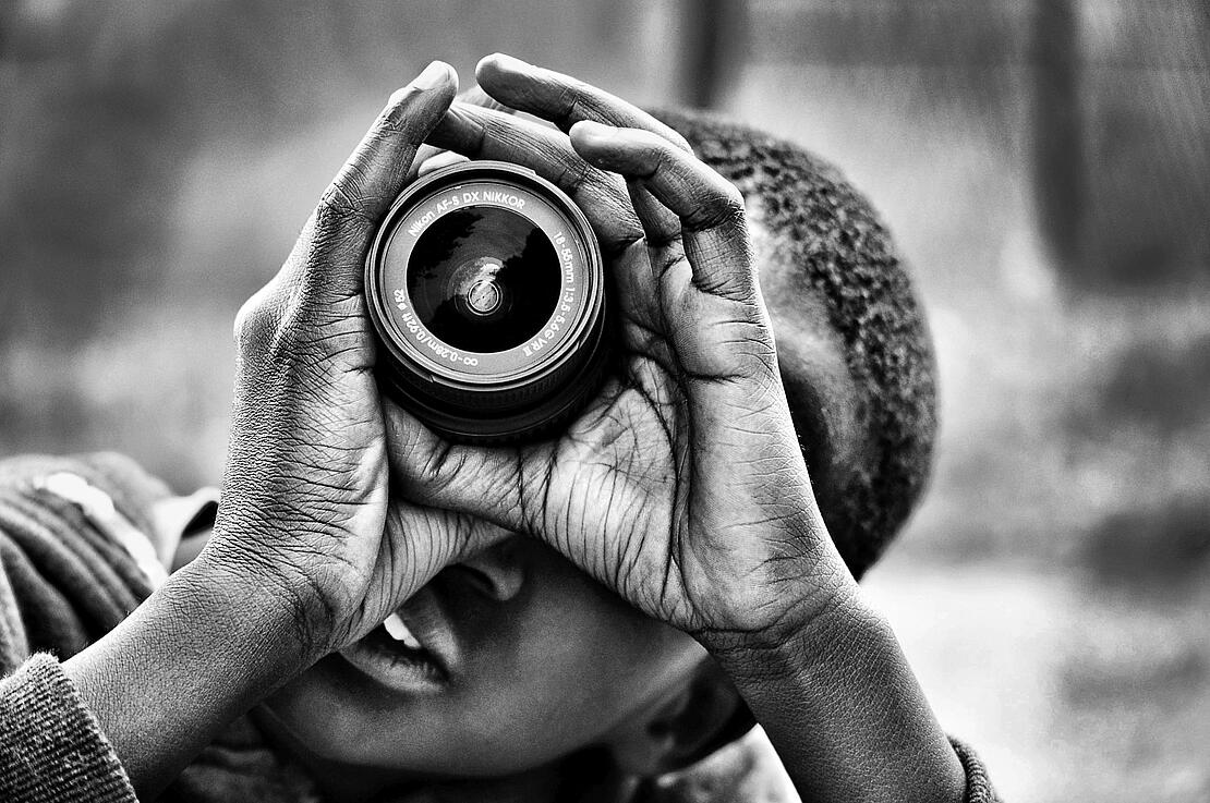 Schwarz-Weiß-Bild zeigt einen farbigen Jungen, der den Betrachter durch das Objektiv einer Kamera anschaut