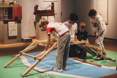 Brückenbau nach Leonardo da Vincis Entwürfen Vier Kinder bauen aus großen Holzelementen eine Brücke.