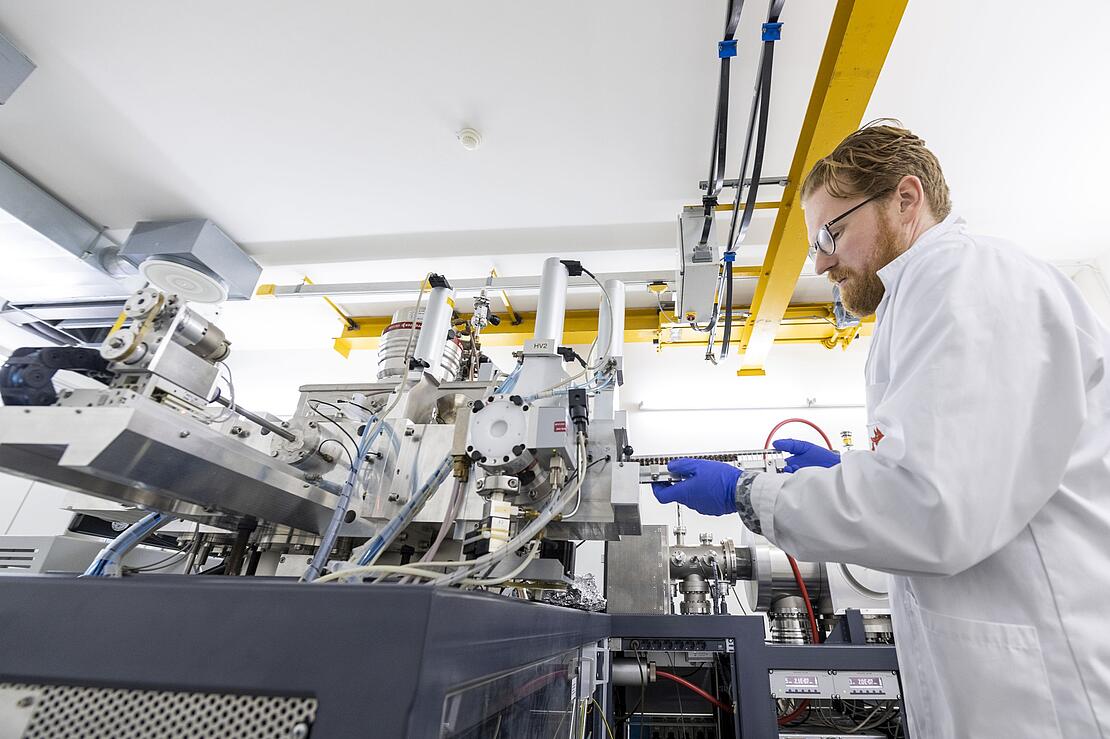 Mann im Laborkittel steht rechts von einem großen kompliziert aussehenden Apparat mit vielen Teilen und Schläuchen. Er scheibt eine Schiene in den Apparat