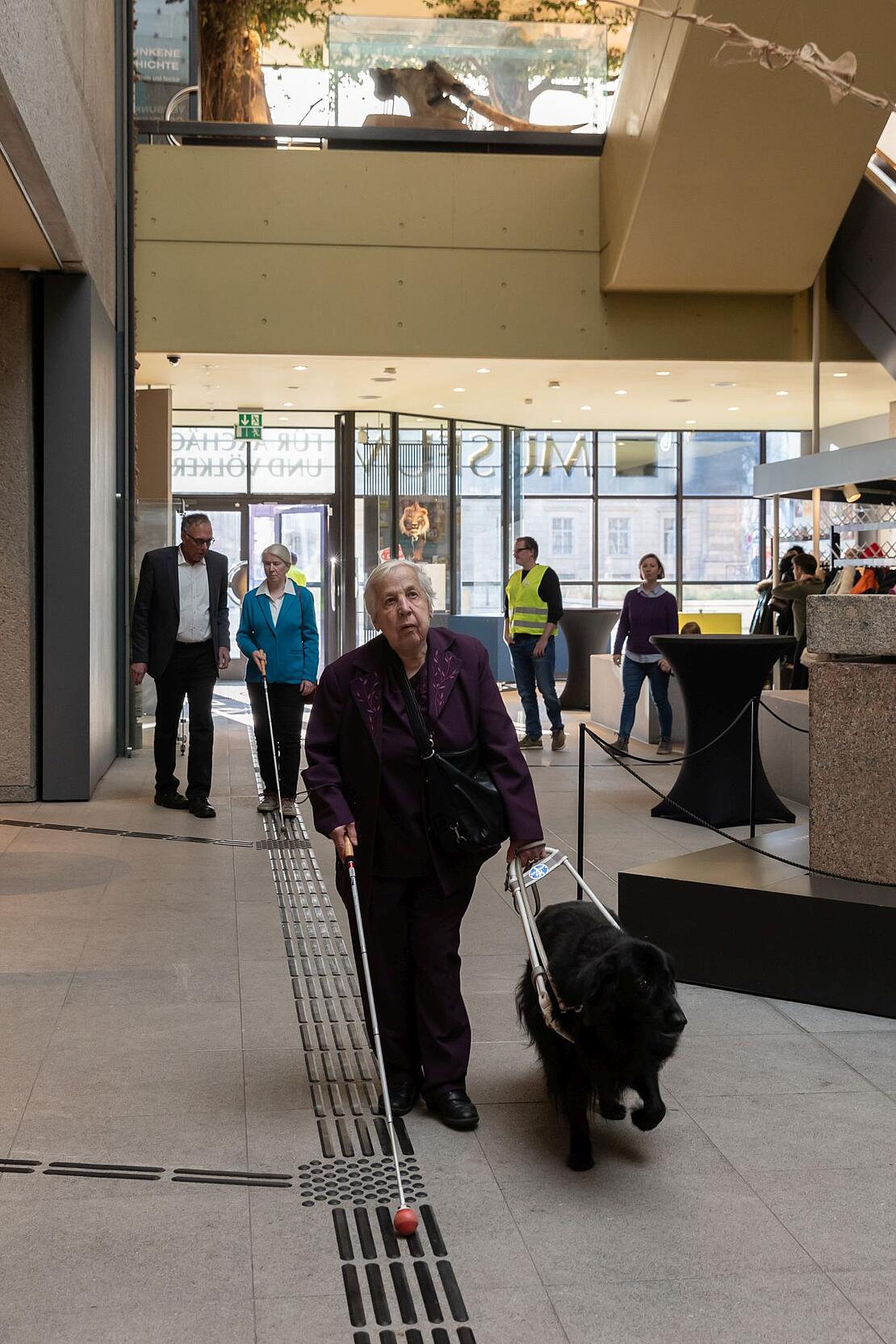Eine Frau mit Hund und Gehstock folgt in einem großen Gebäude einem Blindenleitsystem 