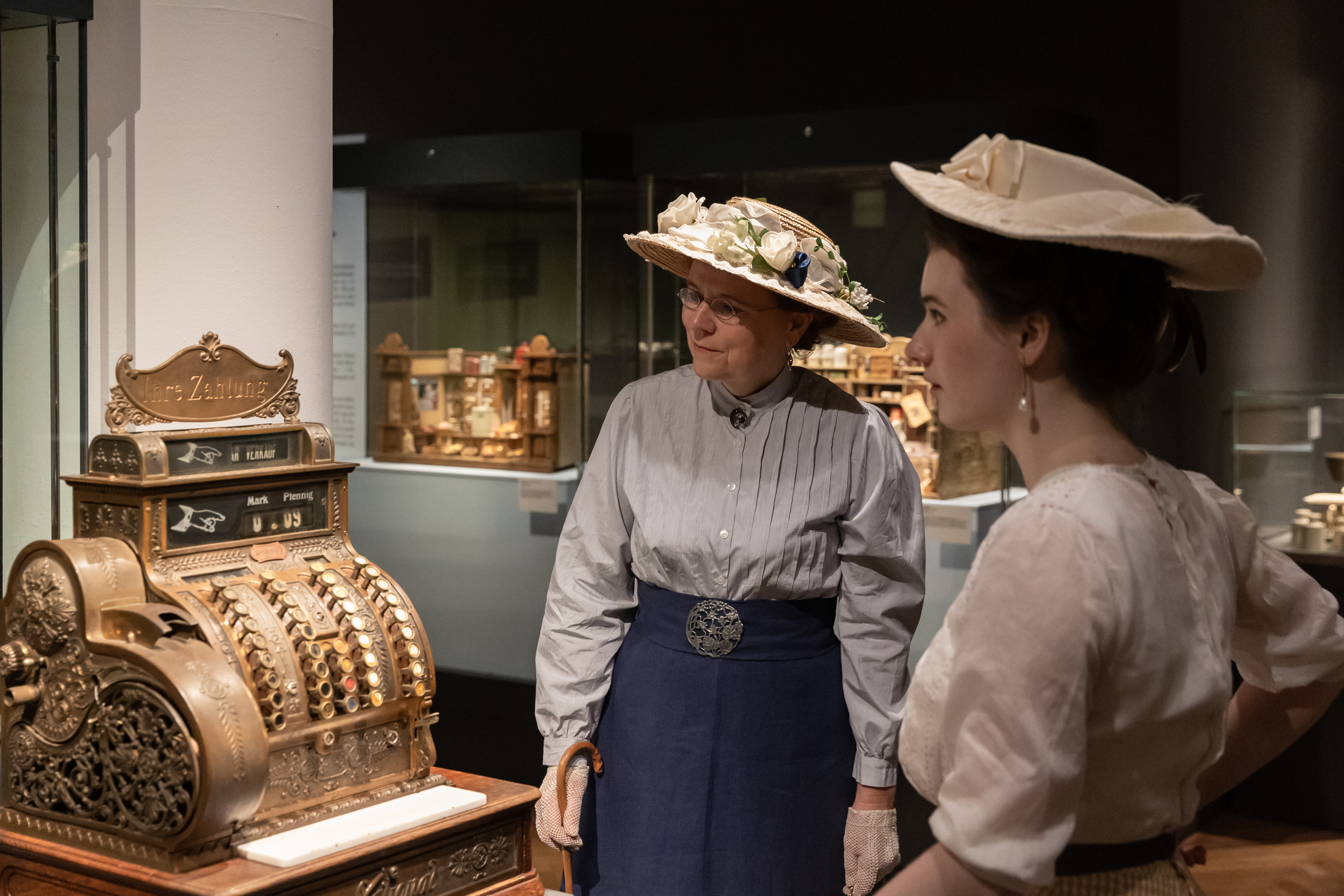 Zwei Besucherinnen im Kostüm betrachten eine Registrierkasse aus dem 19. Jahrhundert